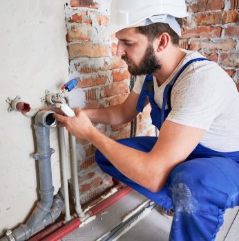 Young plumber, wearing blue uniform and white helmet working with sealant fix of sewer pipe in kitchen or in bathroom in unfinished apartment. Concept of plumbing works and home renovation
